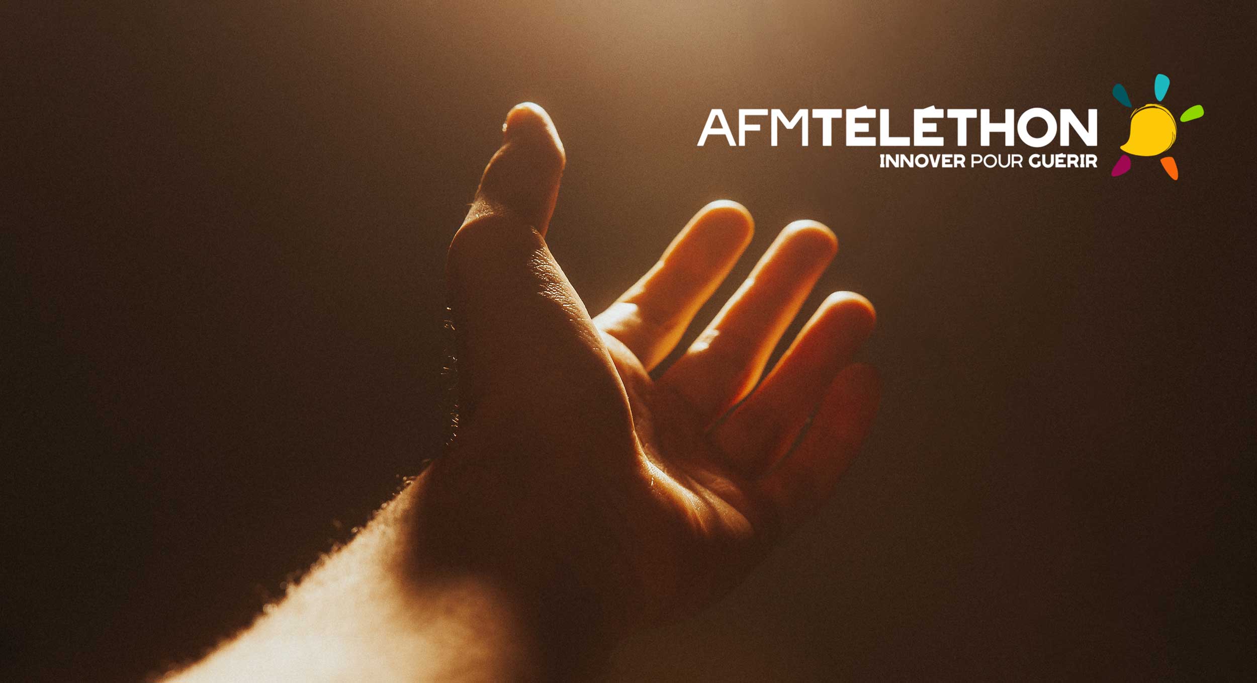 Une main tendu vers le téléthon. Sous une diffuse lumière dorée, une main est ouverte en direction du logo téléthon : symbole de don, douceur et fraternité.
