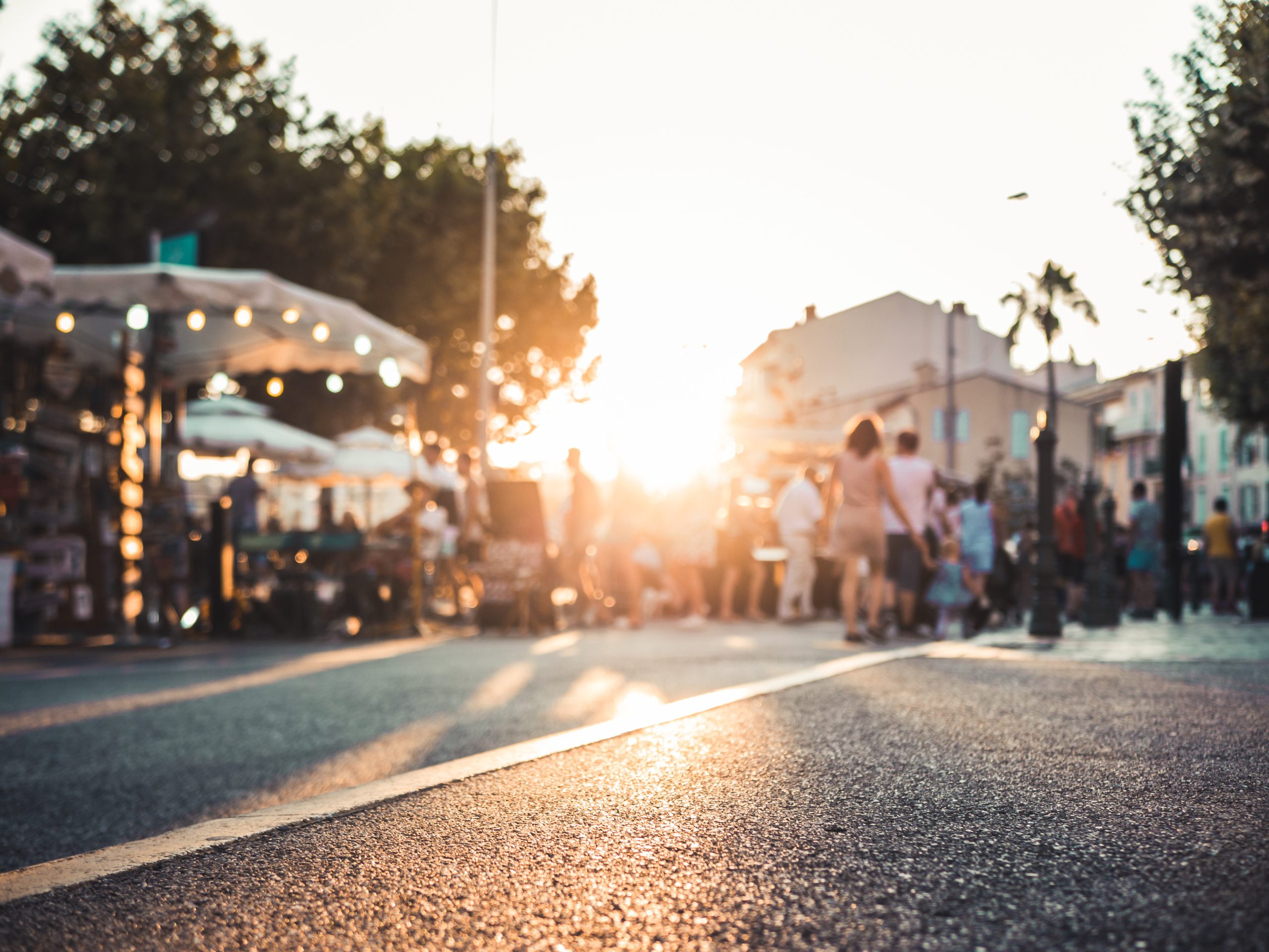 Une vue floue d’un marché en plein air à l'heure du coucher du soleil. Le marché est animé, avec plusieurs tentes et des guirlandes lumineuses créant une ambiance chaleureuse. Des groupes de personnes se déplacent sur une route qui semble être fermée à la circulation, et l'éclairage du coucher du soleil donne une atmosphère agréable à la scène.
