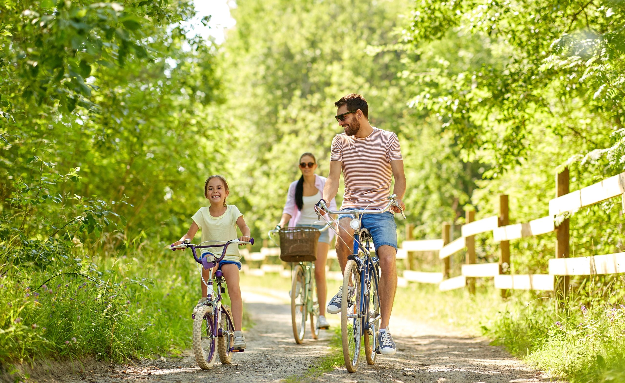 L’image représente une scène en plein air où une famille fait une balade à vélo sur un chemin de terre entouré de verdure. Trois personnes sont visibles : un homme, une femme et une jeune fille. L’homme, positionné à droite de l’image, porte un t-shirt rayé blanc et rouge, un short en jean et des baskets bleues. Il a une barbe et porte des lunettes de soleil. Il roule sur un vélo de type vintage, avec un cadre fin et un guidon courbé. Il affiche un sourire détendu et regarde la jeune fille qui roule à ses côtés. La jeune fille, placée au premier plan à gauche, porte un t-shirt jaune clair et un short bleu. Elle est sur un petit vélo violet et blanc, adapté à son âge, avec des pneus épais. Son expression est joyeuse et enthousiaste, traduisant un moment agréable. Derrière eux, légèrement en retrait, la femme porte une chemise légère ouverte sur un t-shirt blanc, un short en jean et des lunettes de soleil. Elle roule sur un vélo vert pastel équipé d’un panier en osier à l’avant, ce qui renforce l’aspect champêtre et détendu de la scène. Le décor est composé d’un chemin en terre bordé de hautes herbes et d’arbres verdoyants. Une barrière en bois longe la route sur la droite. Le soleil filtre à travers le feuillage, créant une atmosphère lumineuse et estivale. Cette image évoque une sortie familiale agréable en pleine nature.