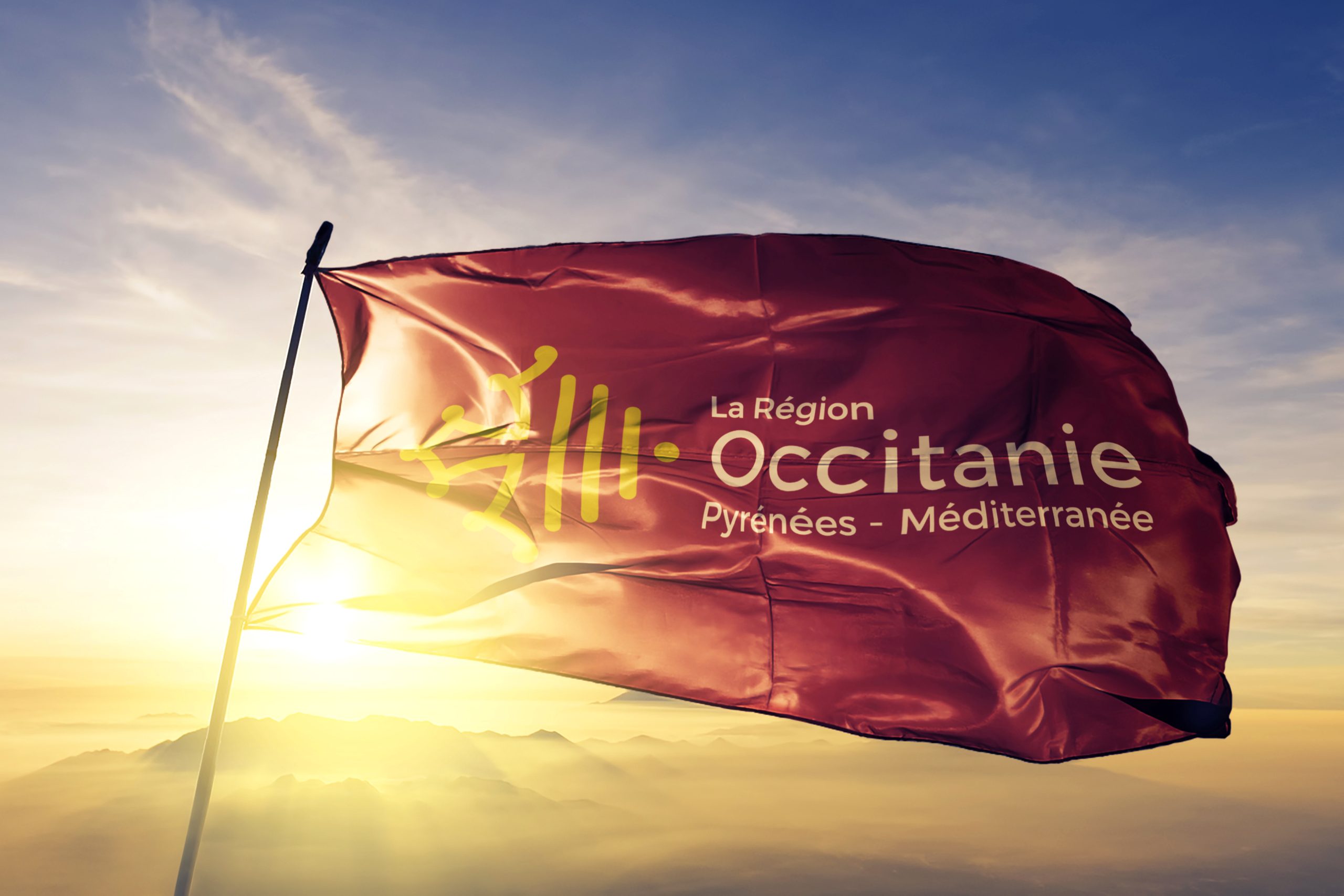 L’image montre un drapeau de la région Occitanie flottant au vent. Le drapeau est principalement de couleur rouge, avec un motif jaune au centre, représentant la croix occitane, un symbole historique et culturel fort. À côté de ce motif, se trouvent les inscriptions "La Région Occitanie Pyrénées - Méditerranée", écrites en lettres blanches et visibles sur la partie inférieure du drapeau. Le logo de la région est également présent, avec des éléments graphiques stylisés représentant la région, accompagnés du nom complet "Occitanie" et du sous-titre "Pyrénées - Méditerranée". Le fond de l’image est composé de couleurs chaudes et naturelles, avec des montagnes visibles sous un ciel lumineux, évoquant un environnement authentique et inspirant. Le drapeau flotte au vent, symbolisant l’identité et la culture de la région. Il n'y a pas de logos ou partenaires supplémentaires visibles dans l’image. L'ensemble de la scène reflète une représentation visuelle de la région Occitanie, mettant en avant son patrimoine et son paysage naturel.
