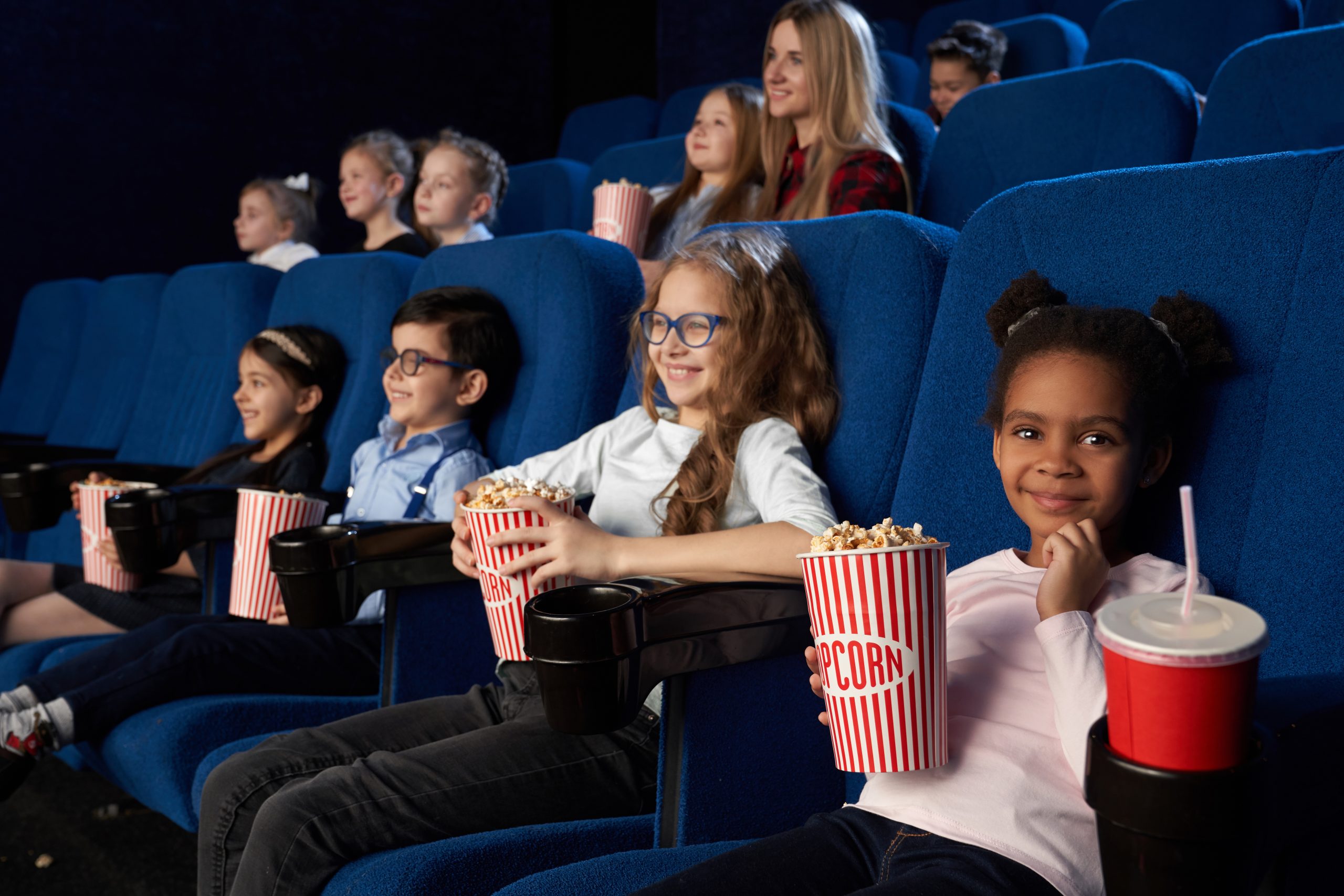 L’image montre un groupe d’enfants et une adulte assis dans une salle de cinéma, profitant d’une projection. Les sièges bleus rembourrés sont alignés en rangées, et chaque enfant tient un gros pot de popcorn rayé rouge et blanc, symbole typique des séances de cinéma. À l’avant-plan, une fillette en pull rose sourit en tenant son popcorn, avec un gobelet de soda rouge avec une paille posé sur l’accoudoir. Les enfants affichent des expressions de plaisir et de concentration, certains portant des lunettes. En arrière-plan, une adulte aux cheveux longs et blonds, vêtue d’une chemise à carreaux rouges, semble accompagner le groupe. L’ambiance est chaleureuse et immersive, capturant l’excitation et la joie d’une sortie au cinéma.