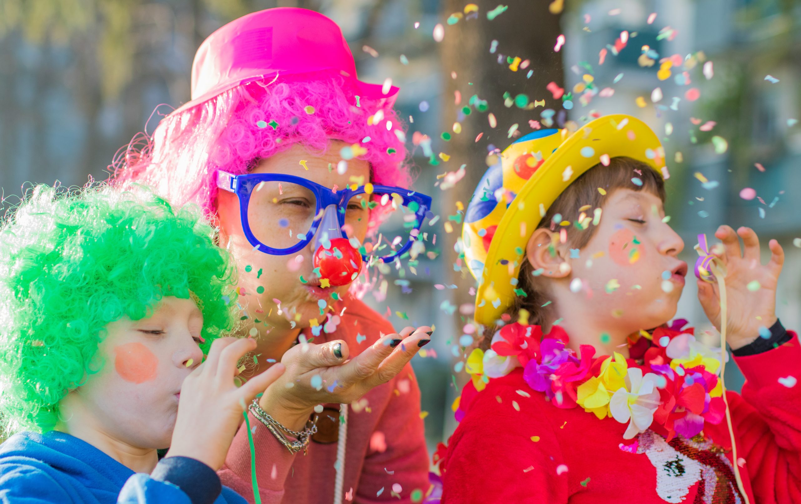 L’image illustre une scène festive en plein air où trois personnes déguisées célèbrent un événement en soufflant des confettis. Au centre, une personne adulte porte une perruque rose bouclée, un chapeau en plastique rose, de grandes lunettes bleues, ainsi qu’un nez rouge de clown. Elle souffle des confettis depuis sa main avec une expression joyeuse. À gauche, un enfant coiffé d’une perruque verte frisée, habillé en bleu, souffle dans un sarbacane, projetant des confettis. Sa joue est maquillée avec une tache rouge. À droite, un autre enfant porte un chapeau jaune décoré de motifs colorés, une écharpe de fleurs hawaïenne autour du cou et un pull rouge avec un motif de licorne. Il souffle également des confettis, les yeux légèrement fermés. Les confettis multicolores flottent dans l’air, renforçant l’ambiance festive. L’arrière-plan est légèrement flou, laissant entrevoir un espace extérieur avec des arbres et des bâtiments. La scène est illuminée par une lumière naturelle, suggérant une célébration en journée.