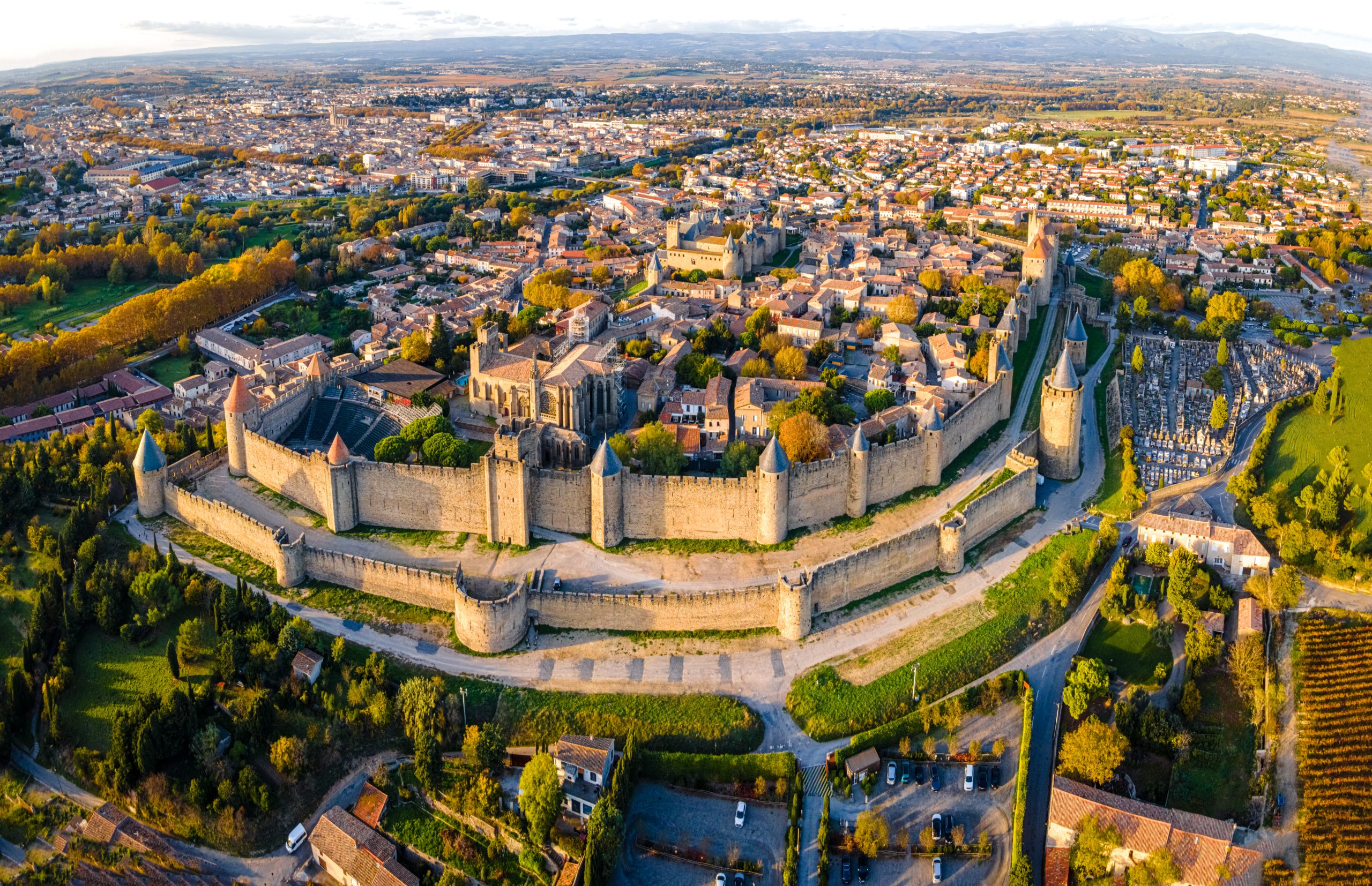 Aerial view of Carcassonne, a French fortified city in France (Vue aérienne de la cité médiévale de Carcassonne) : L’image offre une vue aérienne impressionnante de la cité médiévale de Carcassonne, avec ses remparts bien conservés et ses tours caractéristiques. La ville est entourée par un paysage verdoyant, et les bâtiments à l’intérieur des murailles présentent des toits de tuiles rouges. L’horizon s’étend avec des zones de campagne et quelques éléments urbains, créant un contraste entre l'ancien et le moderne. La lumière dorée du coucher du soleil éclaire la scène, mettant en valeur les détails des murailles et des tours. La structure imposante des fortifications encercle la vieille ville et se détache sur le fond de collines lointaines.