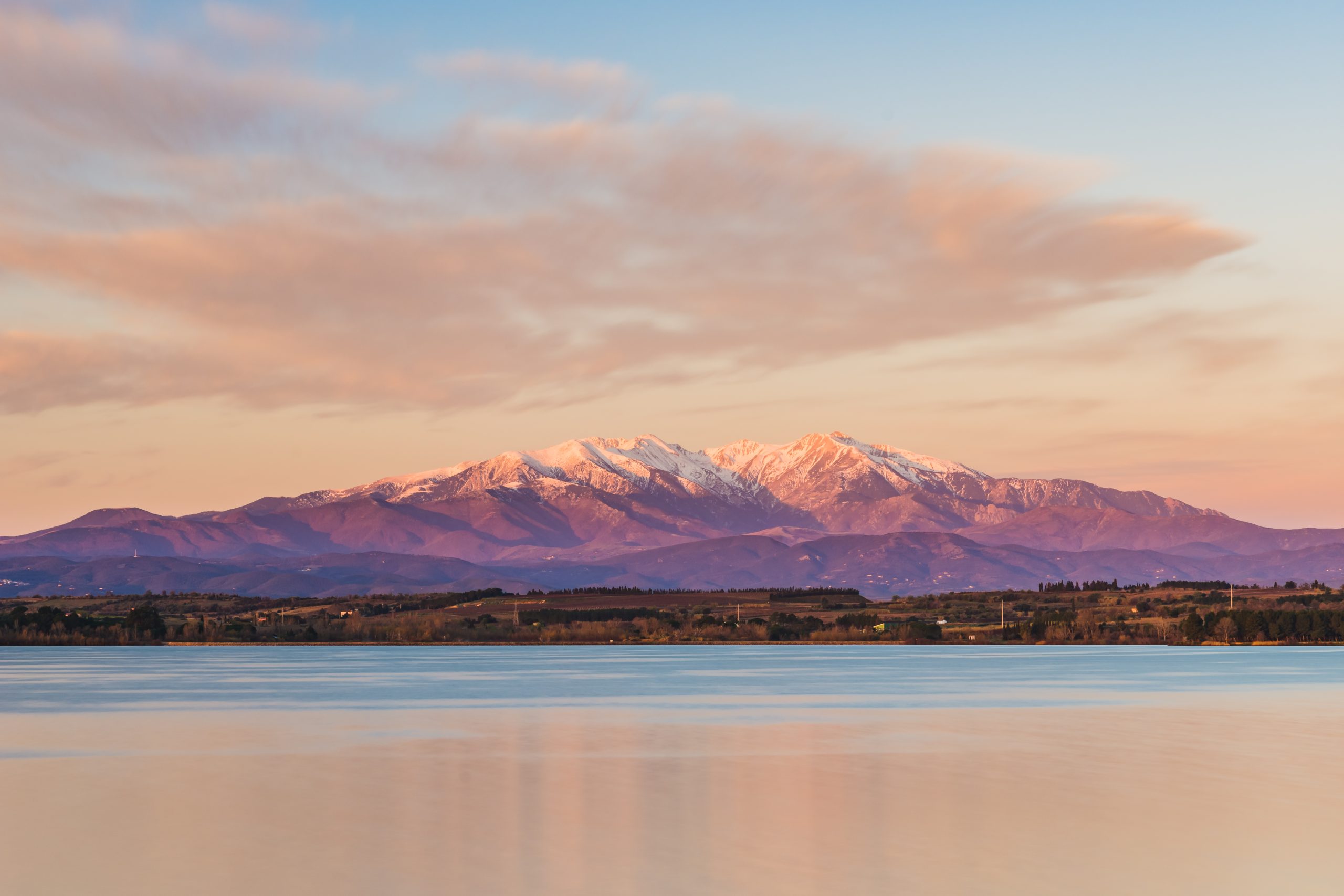 Beautiful sunrise at the lake (View of the Pic du Canigou, France Pyrenees Mountains) (Paysage montagneux au coucher du soleil) : L’image capture un paysage montagnard au coucher du soleil, avec le Canigou recouvert de neige partiellement illuminée par la lumière douce et dorée du soir. L’eau calme d’un lac ou d’un plan d’eau au premier plan reflète partiellement les montagnes, créant un miroir naturel. Le ciel présente des couleurs allant du bleu clair au rose, accentuant la sérénité de la scène. L’image transmet une atmosphère paisible et majestueuse, où les montagnes se dressent majestueusement contre le ciel.