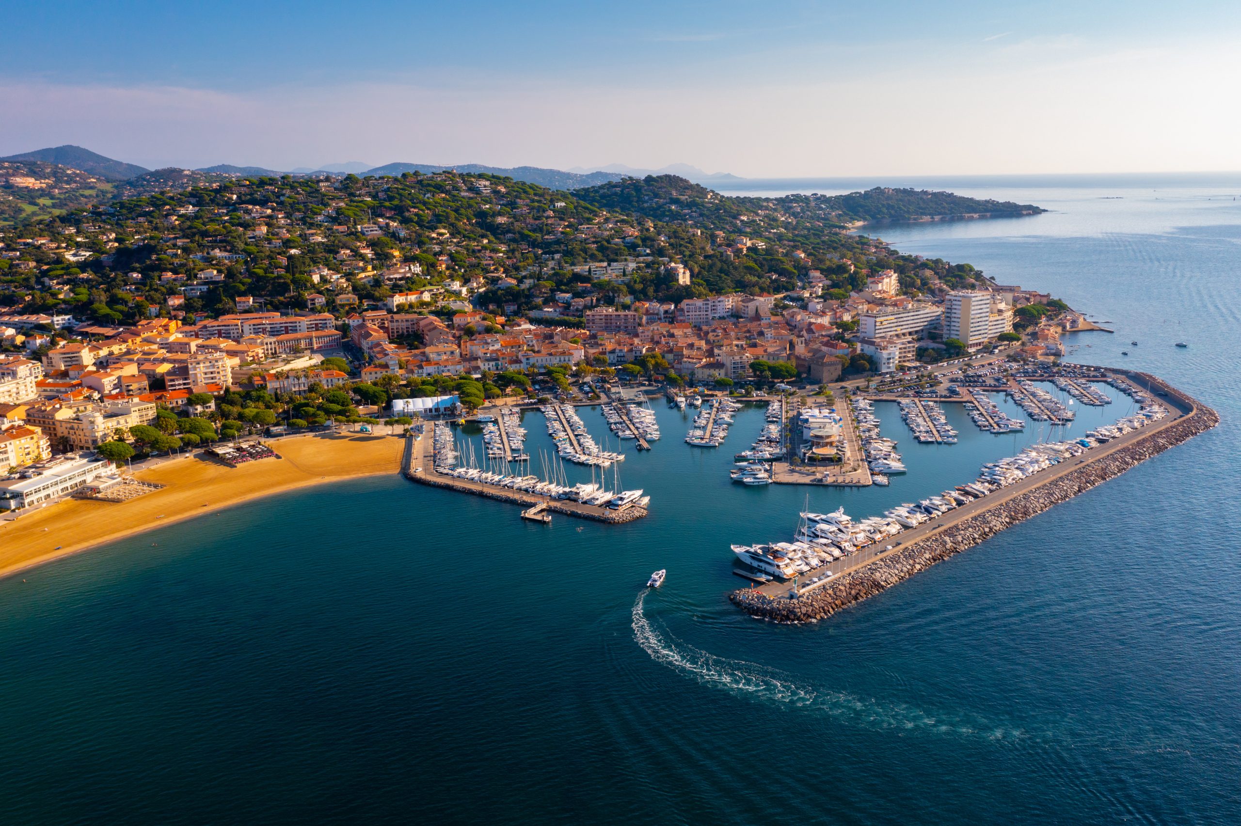 L’image est une vue aérienne d’un port de plaisance situé sur une côte méditerranéenne, bordé par une ville aux toits orangés et entouré de collines verdoyantes. Le port est protégé par deux longues digues en pierre, formant une enceinte où de nombreux bateaux sont amarrés, allant de petits voiliers à de grands yachts. Sur la gauche de l’image, une plage de sable doré s’étend le long du littoral, jouxtant des bâtiments modernes et traditionnels qui composent le centre-ville. Les immeubles sont principalement de faible hauteur, avec une architecture typiquement méditerranéenne, caractérisée par des façades colorées et des toits en tuiles. À l’arrière-plan, le paysage est marqué par des collines couvertes de végétation et parsemées de villas et de résidences privées. La mer, d’un bleu profond, s’étend à l’horizon, reflétant la lumière du soleil sous un ciel dégagé. Un bateau est visible en train de quitter le port, laissant derrière lui une traînée blanche sur l’eau. L’image capture un moment en fin d’après-midi ou en début de soirée, avec une lumière chaude qui accentue les contrastes et met en valeur les couleurs du paysage.