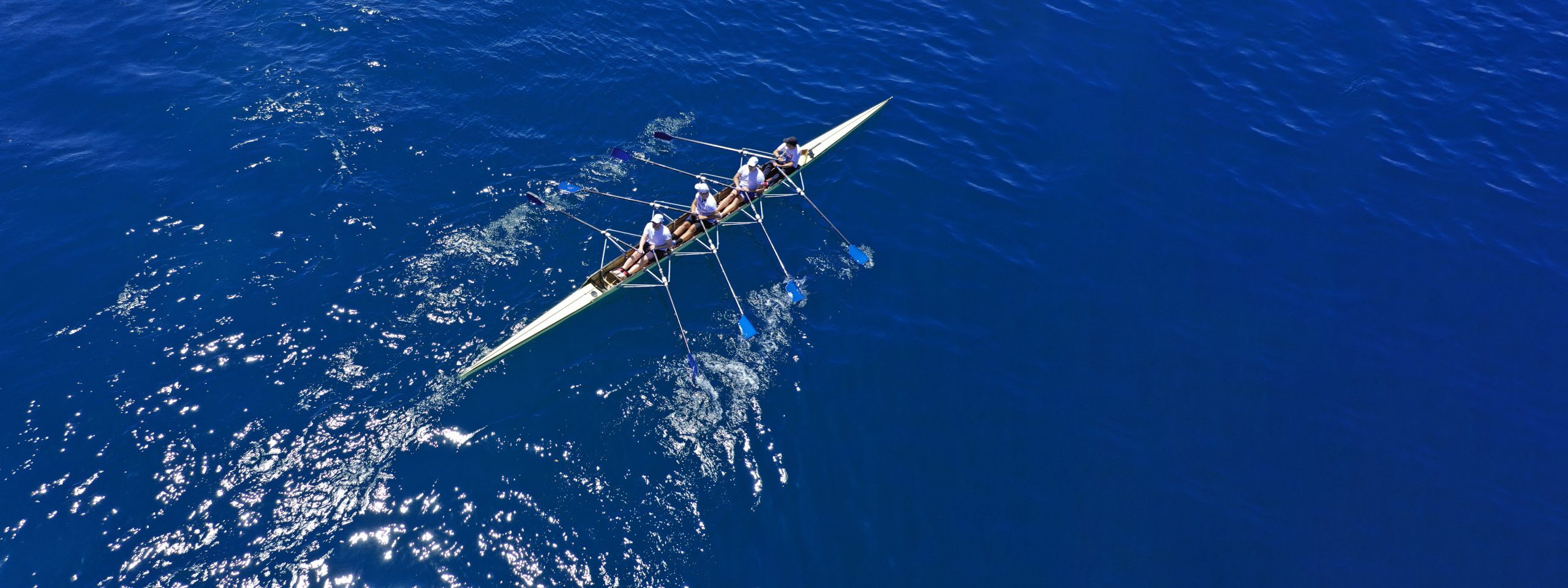 L’image, prise en vue aérienne, montre une équipe d’aviron composée de quatre rameurs synchronisés, vêtus de tenues blanches et de casquettes, naviguant sur une étendue d’eau bleu profond. Leur embarcation est un skiff élancé et profilé, de couleur claire, équipé de rames longues aux pales bleues qui plongent dans l’eau, créant des remous et éclaboussures. La surface de l’eau est légèrement ondulée, avec des reflets du soleil scintillant. L’image capture l’effort collectif et la précision du mouvement, symbolisant la discipline et l’harmonie du sport d’aviron.
