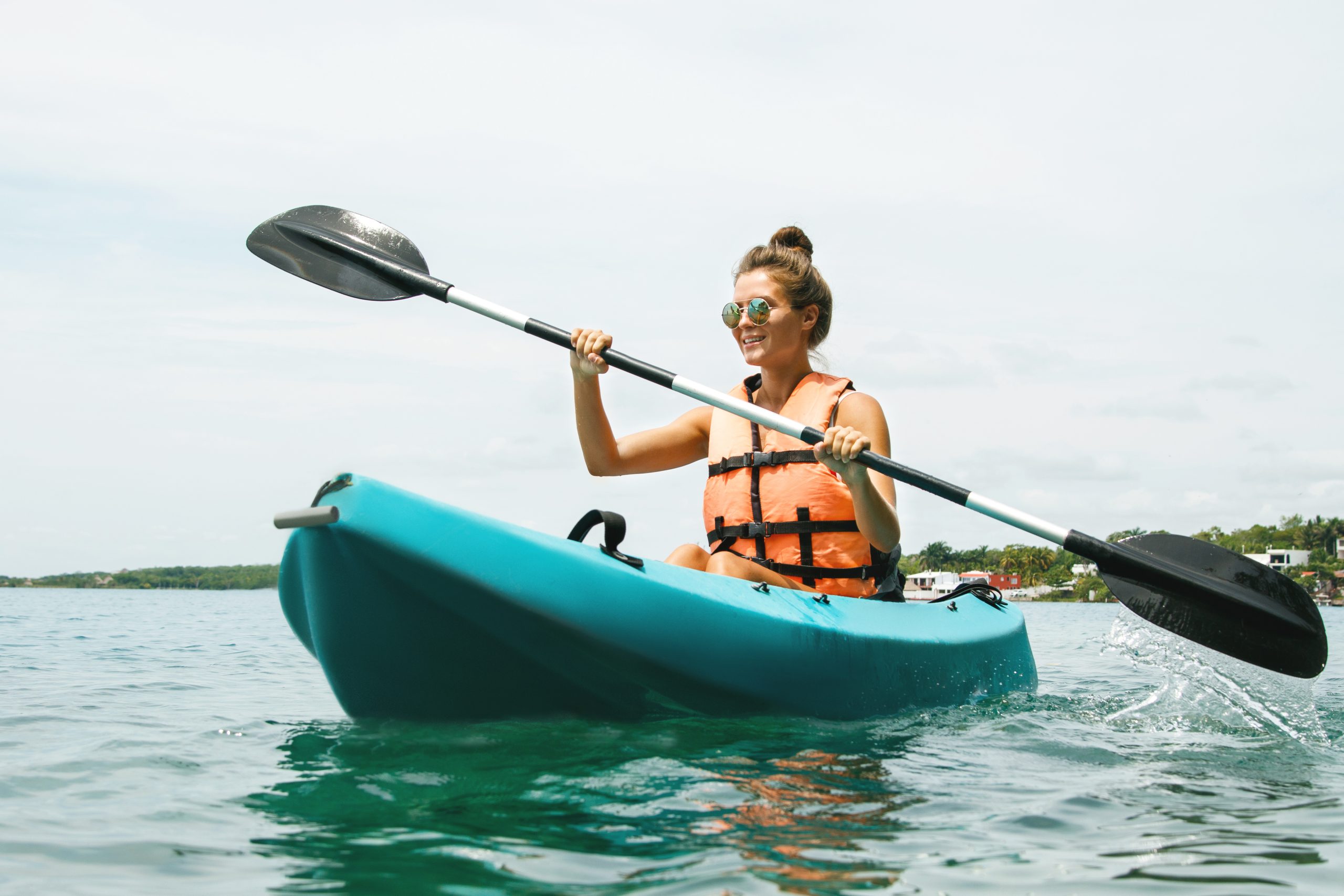 Femme en gilet de sauvetage orange pagayant dans un kayak bleu sur un plan d’eau calme, par temps clair, avec des habitations visibles au loin.