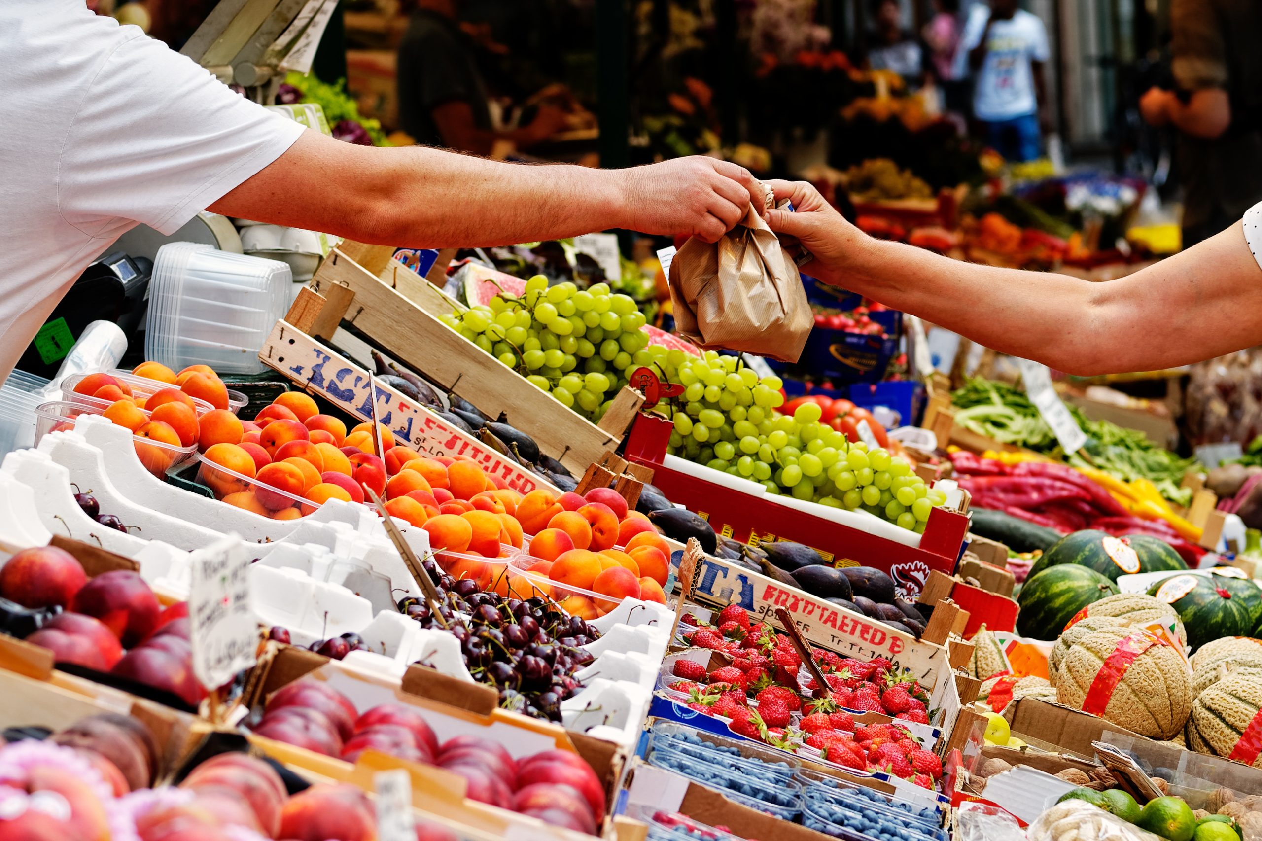 Étal de fruits colorés dans un marché en plein air, avec deux personnes échangeant un sac en papier au-dessus de cagettes remplies de pêches, cerises, fraises, raisins, melons et légumes variés.