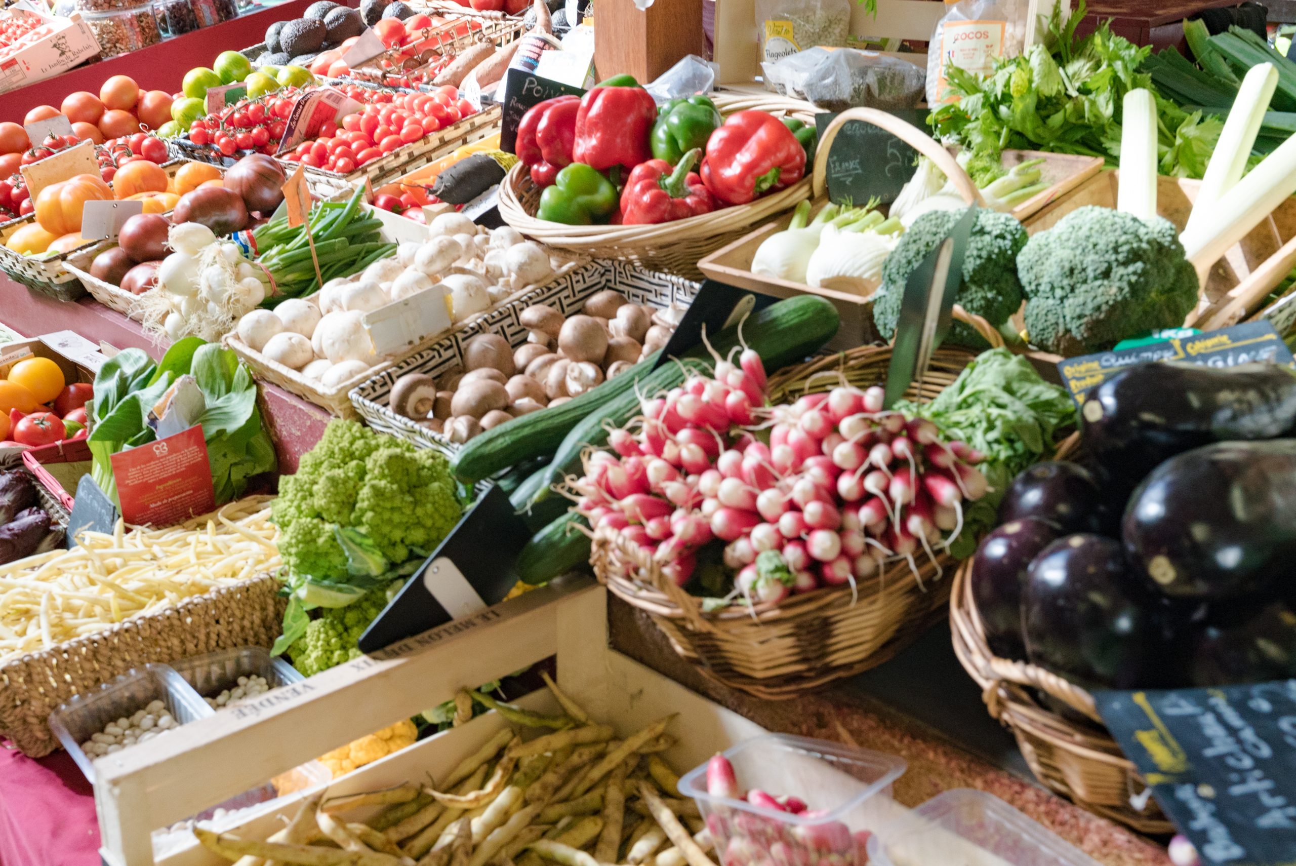 Étal de marché en intérieur avec une grande variété de légumes frais, incluant radis, aubergines, champignons, poivrons, courgettes, brocolis, tomates et haricots, disposés dans des paniers et cagettes.