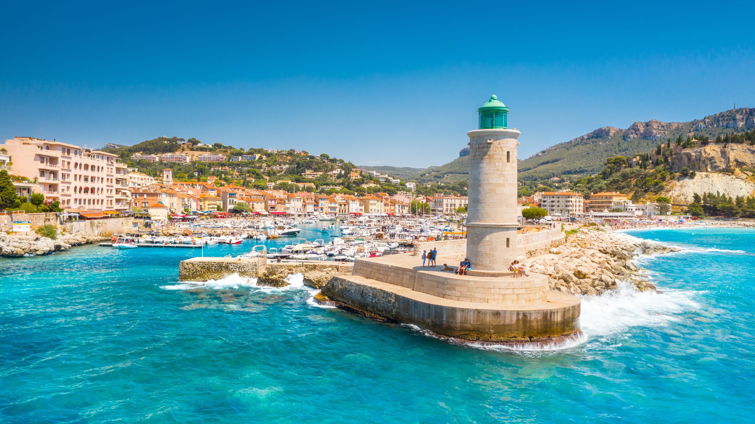 Vue du port de Cassis avec son phare en pierre au premier plan, des bateaux amarrés, des immeubles colorés en bord de mer et des collines verdoyantes en arrière-plan sous un ciel bleu clair.