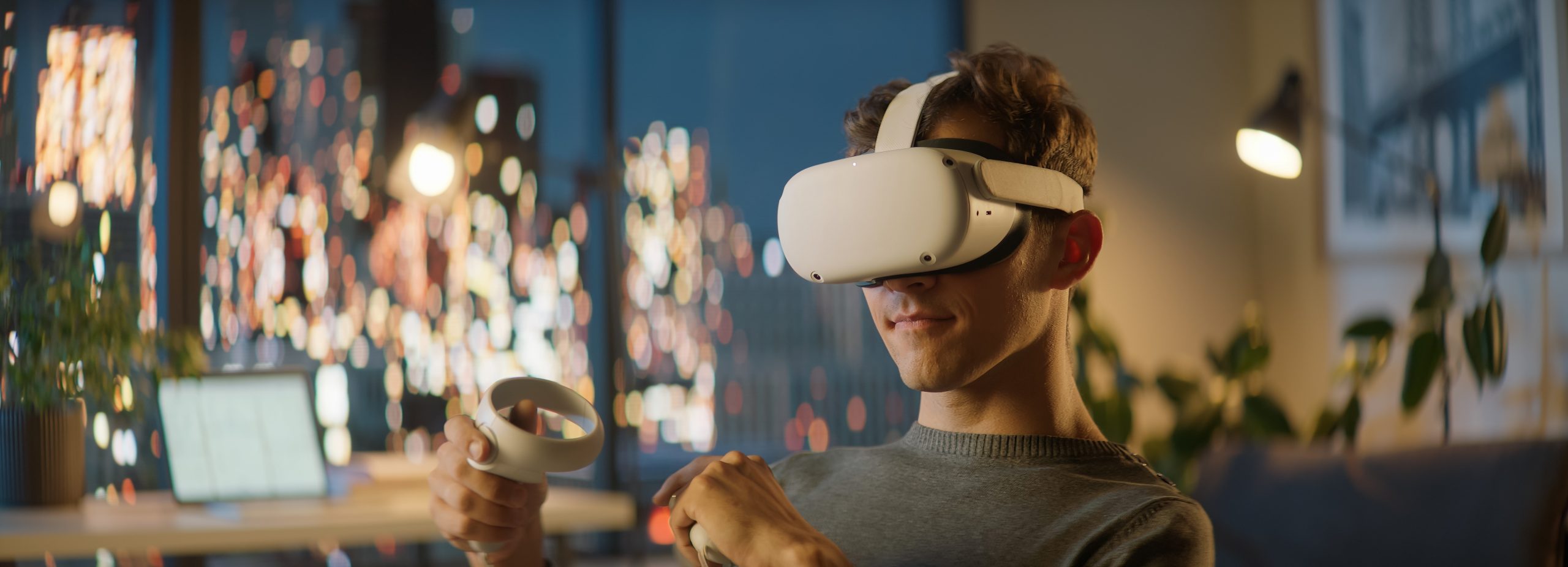 Jeune homme utilisant un casque de réalité virtuelle avec des manettes, assis dans un intérieur moderne en soirée, avec des lumières floues de la ville en arrière-plan.