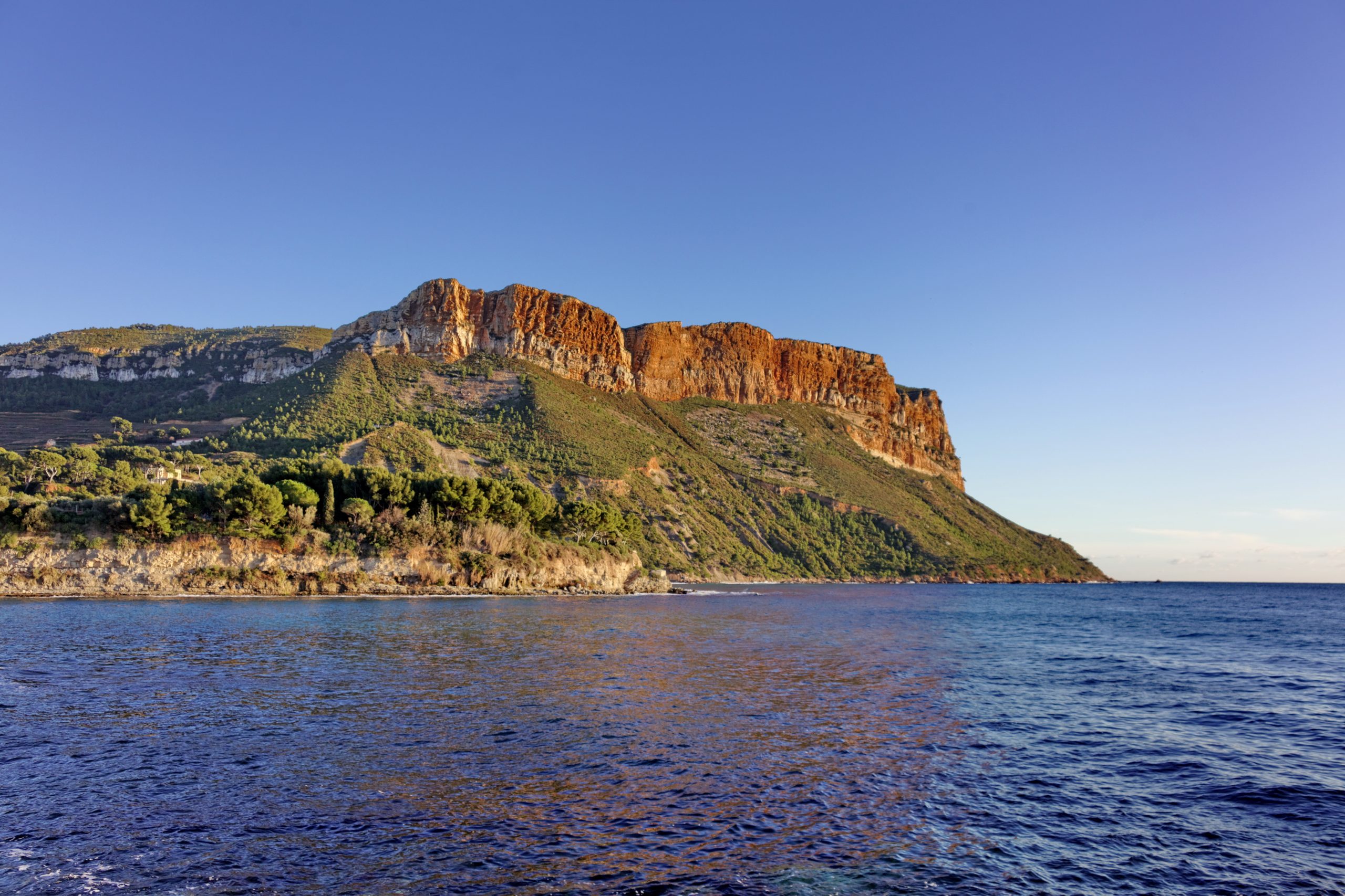 Vue côtière sur le Cap Canaille, grande falaise rocheuse aux teintes ocres dominant la mer Méditerranée, avec végétation sur les pentes et ciel dégagé en arrière-plan.